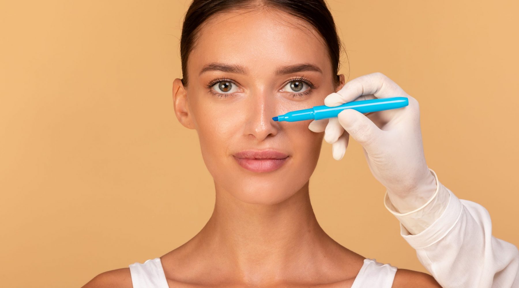 Rhinoplasty concept. Doctor making marks on patient's face, young caucasian lady on consultation at surgeon, standing on beige background. Facial plastic surgery concept